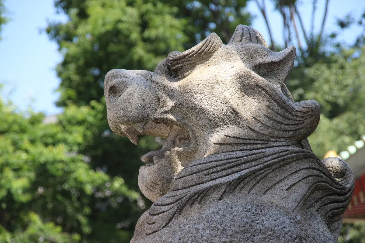 A weathered stone guardian lion with open mouth set against green trees at Kanda Myojin shrine